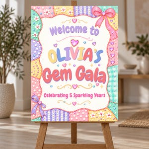 Può includere: Un cartello rettangolare su un cavalletto in legno con un bordo patchwork color pastello decorato con fiocchi, fiori e gemme. Il testo recita: Welcome to OLIVIA'S Gem Gala Celebrating 5 Sparkling Years. Include gemme a forma di cuore.