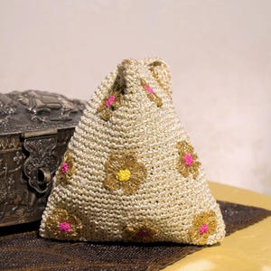 May include: A triangular-shaped crochet pouch in a metallic gold yarn. The surface features embroidered floral motifs with gold petals and centers in bright pink or yellow. The pouch has a textured, woven appearance with a small handle at the top.