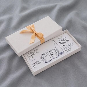 Puede incluir: Una caja de regalo rectangular blanca con una cinta dorada, que contiene una tarjeta impresa titulada Pottery & Prosecco for Two. La tarjeta presenta una ilustración dibujada a mano de dos tazas y una botella, con el texto A Christmas present that doesn't need dusting.