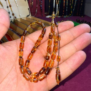 May include: A prayer bead strand featuring translucent, amber-coloured cylindrical beads with dark brown marbled patterns. The beads are strung on a thin black cord, ending in a decorative, elongated terminal bead with a matching amber and brown finish.