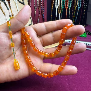 May include: A prayer bead strand featuring translucent orange cylindrical beads strung on a green cord. The strand includes a decorative tassel component with a small silver-toned circular accent and a larger cylindrical orange bead at the end.