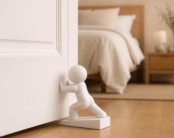 May include: A white doorstop shaped like a stylized human figure in a pushing pose. The figure has a smooth, rounded head and body, with its hands pressed against the surface of a white door to hold it open, mounted on a wedge-shaped base.