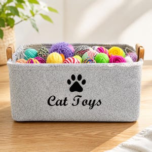 Op de afbeelding: Een rechthoekige opbergmand van lichtgrijs vilt met twee houten handvatten. De voorzijde is voorzien van een zwarte pootafdruk en de tekst "Cat Toys" in een cursief lettertype. De mand is gevuld met diverse kleurrijke bollen wol en speeltjes.