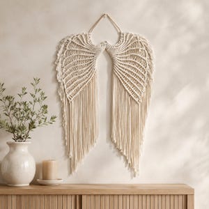May include: A cream-coloured macrame wall hanging shaped like a pair of angel wings. The piece is constructed from knotted cotton cord, featuring intricate fan-like patterns at the top that transition into long, straight, flowing fringe at the bottom.
