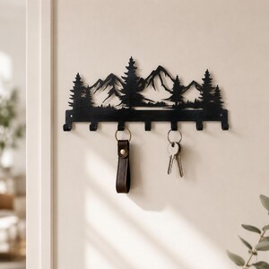 May include: A black metal wall-mounted key holder featuring a silhouette of a mountain range with pine trees. The horizontal rack includes seven downward-facing hooks for hanging items, with two sets of keys currently suspended from the hooks.
