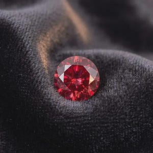 May include: A round, faceted gemstone with a deep, vibrant red colour. The stone features a brilliant cut with numerous triangular and kite-shaped facets that reflect light, creating a complex pattern of dark and bright red tones across its surface.