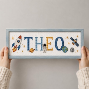 Può includere: Un ricamo a punto croce incorniciato che riporta il nome THEO in grandi lettere blu e gialle. Il disegno include un razzo, un piccolo astronauta, un pianeta con anelli, un globo, un satellite e piccole stelle su uno sfondo di tessuto bianco.