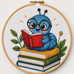 May include: A cross-stitch embroidery in a round wooden hoop featuring a blue caterpillar with large black glasses reading an open red book. The caterpillar sits atop a stack of three books in yellow, green, and white, surrounded by green leaves.