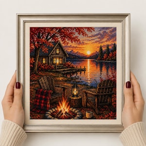 Op de afbeelding: Een ingelijst kruissteek kunstwerk met een blokhut aan een meer bij zonsondergang. De scène toont twee houten stoelen, een gloeiend kampvuur, een lantaarn en een rood geruite deken. De oranje en paarse lucht reflecteert in het wateroppervlak.