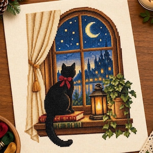 May include: A cross-stitch embroidery on white fabric depicting a black cat with a red bow sitting on a book by a window. The scene shows a night view with a crescent moon, stars, a city silhouette, a lit lantern, and a green potted plant.