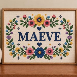 以下が含まれることがあります： オフホワイトの布地にダークブルーの大文字で「MAEVE」と刺繍された、額装済みのクロスステッチ作品。文字の周囲は、黄色、ピンク、青の花々、緑の葉、そして小さなダークブルーのハートで構成されたカラフルな花輪で囲まれています。