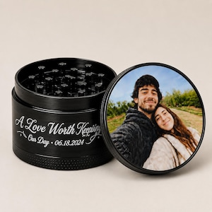 Puede incluir: Un molinillo de hierbas cilíndrico negro con una foto personalizada de una pareja en la tapa. El lateral presenta el texto grabado A Love Worth Keeping Our Day 06.18.2024. El interior contiene múltiples dientes de metal negro para moler hierbas.