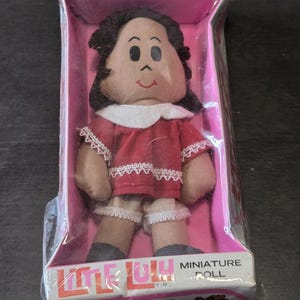 May include: A Little Lulu miniature doll with dark hair, a red dress with white lace trim, and white bloomers, packaged in a pink and white box. The box features the text Everyone's favorite little girl and Little Lulu Miniature Doll.