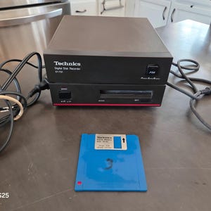 May include: A black Technics Digital Disk Recorder SY-FD1 with a power cord and a separate blue floppy disk. The device features a front-facing power button, a disk slot, and a small square button labeled F30. The blue disk has a Technics label.