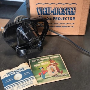 May include: A black View-Master Junior Projector with its original cardboard box, a View-Master reel, and a Huckleberry Hound and Yogi Bear paper sleeve. The projector has a lens and a power cord. The box features blue text and wavy lines.