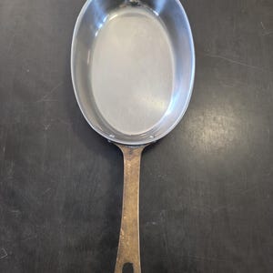 May include: An oval-shaped metal pan with a smooth, reflective silver-toned interior and a long, weathered brass-colored handle. The handle features a small, elongated hole at the end for hanging, and the pan is attached to the handle with rivets.