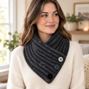 Puede incluir: Una bufanda tipo cuello de punto acanalado en color gris carbón oscuro con un cierre frontal diagonal superpuesto, asegurado por tres botones grandes y redondos en tonos gris y negro, sobre un suéter de punto de manga larga color crema.