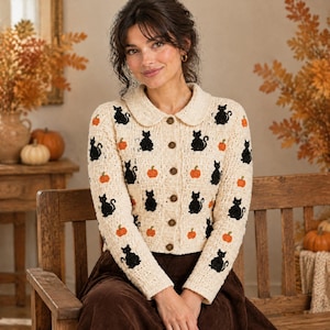May include: A cream-colored long-sleeve cardigan featuring a repeating pattern of embroidered black cats and orange pumpkins. The knit garment has a rounded collar, a button-down front with brown buttons, and a textured, woven fabric construction.