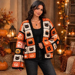 May include: A long-sleeved crochet cardigan featuring a patchwork pattern of black cats, orange cauldrons, and jack-o'-lanterns in a colour palette of black, orange, and cream. The garment has an open front design with wide sleeves and a square-neck motif.