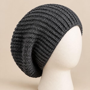 May include: A dark charcoal gray slouchy beanie hat featuring a prominent vertical ribbed knit texture. The hat is constructed from a thick yarn material and displays a relaxed, rounded shape that drapes slightly toward the back of the head.