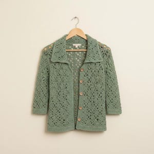 May include: A sage green crochet cardigan featuring an openwork lace pattern, a collared neckline, and three-quarter length sleeves. The garment is fastened with five small, round, light-coloured buttons down the centre front and hangs on a hanger.