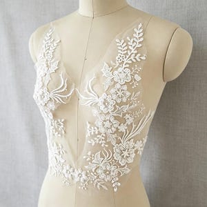 May include: A white floral lace appliqué featuring intricate embroidered flowers, leaves, and vines on a sheer mesh base. The design is arranged in a symmetrical pattern with a deep V-shaped neckline, showcasing detailed stitching and delicate textures.
