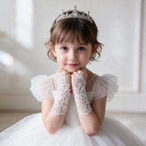 Puede incluir: Una tiara plateada con intrincados detalles metálicos y cristales transparentes, combinada con guantes de encaje blanco sin dedos adornados con delicados bordados florales y pequeños diamantes de imitación brillantes que cubren los antebrazos.