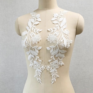 May include: A pair of white lace appliques featuring intricate floral and leaf embroidery on a sheer mesh base. The symmetrical design displays detailed petals, stems, and foliage, creating a delicate pattern intended for garment embellishment.