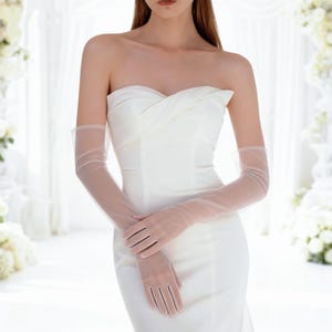 May include: A strapless white gown featuring a fitted bodice with a subtle twist detail at the chest, paired with sheer, elbow-length opera gloves. The dress has a clean, smooth silhouette that extends past the waistline, creating a formal look.