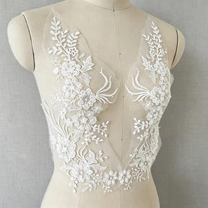 Può includere: Un'applicazione in pizzo floreale bianco con intricati fiori ricamati e tralci di foglie su una base in rete trasparente. Il design è simmetrico, disposto in due pannelli verticali che seguono la forma di una profonda scollatura a V.