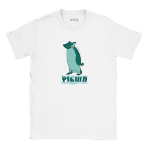 May include: A white short-sleeve t-shirt featuring a central graphic of a teal pig-penguin hybrid creature standing on a small patch of ice. Below the creature, the text PIGUIN is printed in a bold, dark teal font. The shirt has a crew neckline.
