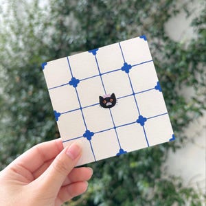 May include: A square off-white card featuring a blue grid pattern with small blue cross shapes at each intersection. Centered on the card is a small illustration of a black cat with orange eyes, a light pink bow, and a grey muzzle.