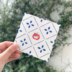 May include: A square white card featuring a blue diamond grid pattern with small blue floral accents in each section. A single illustration of a cat wearing a red apple-shaped hat is centred within one of the blue diamond grid sections.