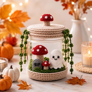 Op de afbeelding: Een doorzichtige glazen pot met een beige gehaakt deksel bekroond met een rood-witte paddenstoel. Binnenin staan een gehaakt wit spookje, een grote rode paddenstoel, een kleine rode paddenstoel en een grijze grafsteen met de tekst RIP.