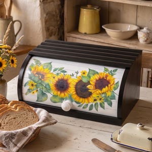 May include: A black wooden bread box with a roll-top door featuring a printed sunflower design. The front panel displays yellow sunflowers with green leaves on a white background and a white knob. The bread box sits on a white wooden table.