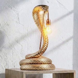 May include: A gold-coloured table lamp shaped like a coiled cobra snake. The lamp features a textured, scale-like surface throughout its body. A single exposed light bulb hangs from the cobra's hood, emitting a warm glow from the fixture.