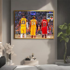 May include: A rectangular wall art print featuring three basketball players from behind, standing on a court. The players wear jerseys with the names James 23, Bryant 24, and Jordan 23. The print has a gold frame and depicts a crowded arena background.
