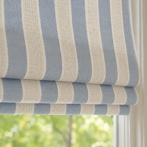 Könnte beinhalten: Ein Faltrollo mit vertikalem Streifenmuster in Hellblau und Off-White. Der Stoff hat eine sichtbare Webstruktur mit Fischgrätenmuster. Das Rollo ist unten teilweise gefaltet, wodurch horizontale Falten über dem Fenster entstehen.