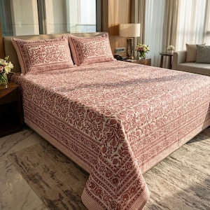May include: A bedspread and two matching pillowcases featuring a repeating floral and pomegranate pattern in muted red on a cream background. The textile has a wide decorative border with geometric and botanical motifs along the edges.