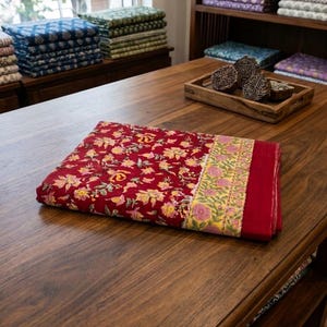 May include: A folded rectangular textile featuring a deep red base with a multi-colored floral pattern in yellow, pink, and green. The fabric includes a distinct yellow border with pink floral motifs and a solid red edge on the right side.