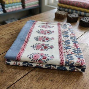 Op de afbeelding: Een gevouwen textiel met een crèmekleurig middenpaneel met een herhalend patroon van roze en blauwe bloemmotieven. De stof is omrand door een effen lichtblauwe strook en een decoratieve band met blauwe bladeren en roze bloemen aan de randen.