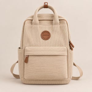 Può includere: Uno zaino in velluto a coste beige con una tasca frontale con cerniera, due manici superiori con chiusura a scatto in similpelle marrone e una toppa circolare marrone sul davanti con il testo YISHUIHAN. Include tasche laterali e spallacci.