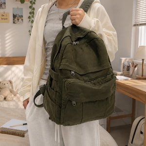 Puede incluir: Una mochila de lona en color verde oliva oscuro que cuenta con un compartimento principal grande, un bolsillo frontal secundario y un bolsillo inferior más pequeño, todos con cremalleras metálicas plateadas, asa superior y correas.