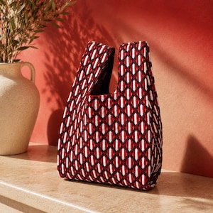 Peut inclure: Un sac fourre-tout matelassé présentant un motif géométrique répétitif en losanges rouge foncé, blanc et noir. Le sac possède des poignées intégrées, une silhouette structurée et une surface en tissu texturé créant une profondeur visuelle.