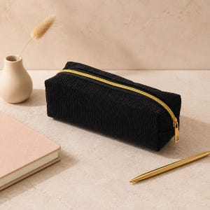 Peut inclure: Une trousse à crayons rectangulaire en velours côtelé noir dotée d'une fermeture éclair en métal doré sur le dessus. Le tissu présente une texture côtelée verticale distincte et la tirette est une languette métallique rectangulaire.