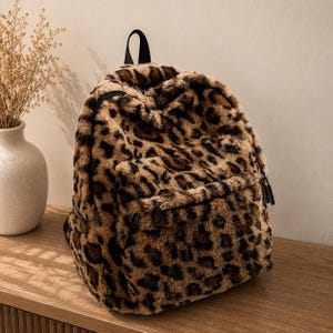 Peut inclure: Un sac à dos en fausse fourrure douce avec un motif léopard composé de taches brunes et noires. Le sac présente un compartiment principal zippé, une poche frontale zippée plus petite et une poignée supérieure noire pour le transport.