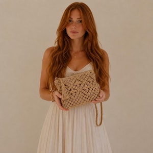 May include: A beige macrame clutch bag featuring a geometric diamond pattern, a decorative tassel on the side, and a matching braided wrist strap. The bag is constructed from woven cord with a textured, knotted design throughout the exterior.
