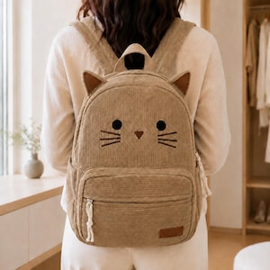 Puede incluir: Una mochila de pana color canela con un diseño de cara de gato que incluye dos orejas puntiagudas, ojos negros circulares bordados, una pequeña nariz triangular y bigotes. La bolsa cuenta con un bolsillo frontal con cremallera y tirador.
