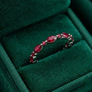 Puede incluir: Un anillo de tono plateado que presenta una serie de rubíes de color rojo intenso, alternando cortes marquesa y redondos a lo largo de la banda. Las piedras están dispuestas en un patrón repetitivo, mostrando un tono carmesí vibrante sobre el metal.