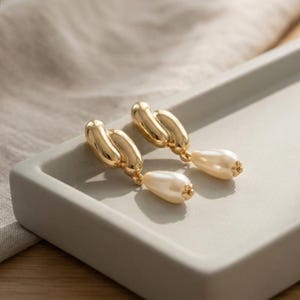 Puede incluir: Un par de pendientes dorados con una parte superior de metal pulido y retorcido, conectada a un colgante de perla sintética de color crema con forma de lágrima. Los pendientes tienen un acabado metálico reflectante y un diseño clásico.
